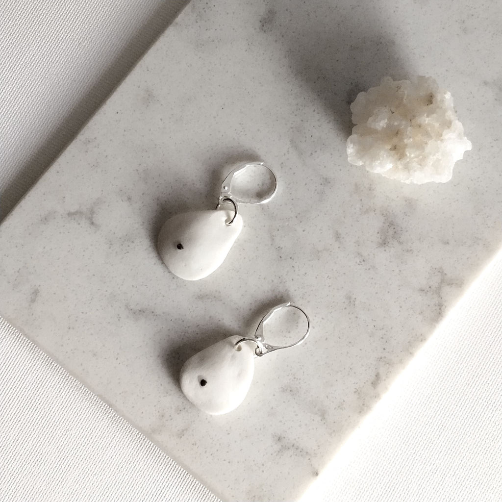 Orecchini Soffio in porcellana bianca con inclusioni di platino e monachelle in argento, posa dall’alto su superficie chiara.