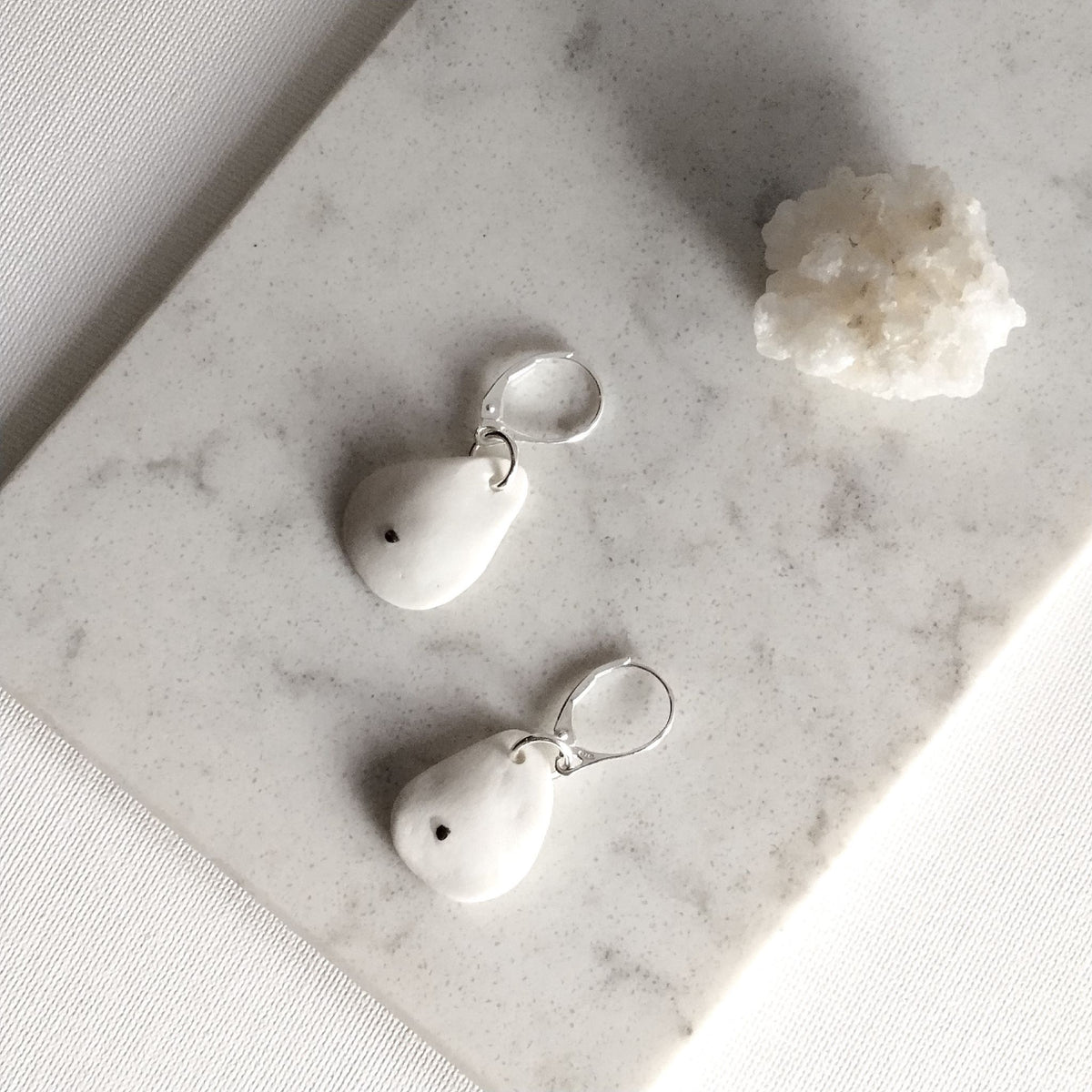 Orecchini Soffio in porcellana bianca con inclusioni di platino e monachelle in argento, posa dall’alto su superficie chiara.