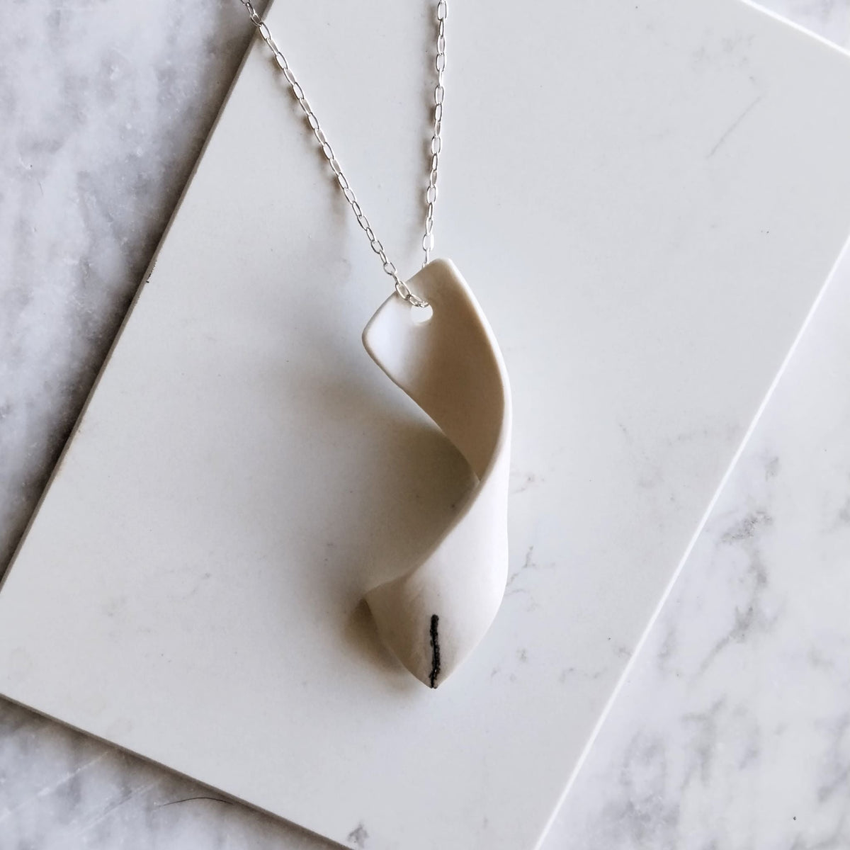 Collana Turbine Maviart in posa dall’alto su marmo bianco, con pendente sinuoso in porcellana bianca e catena in argento.