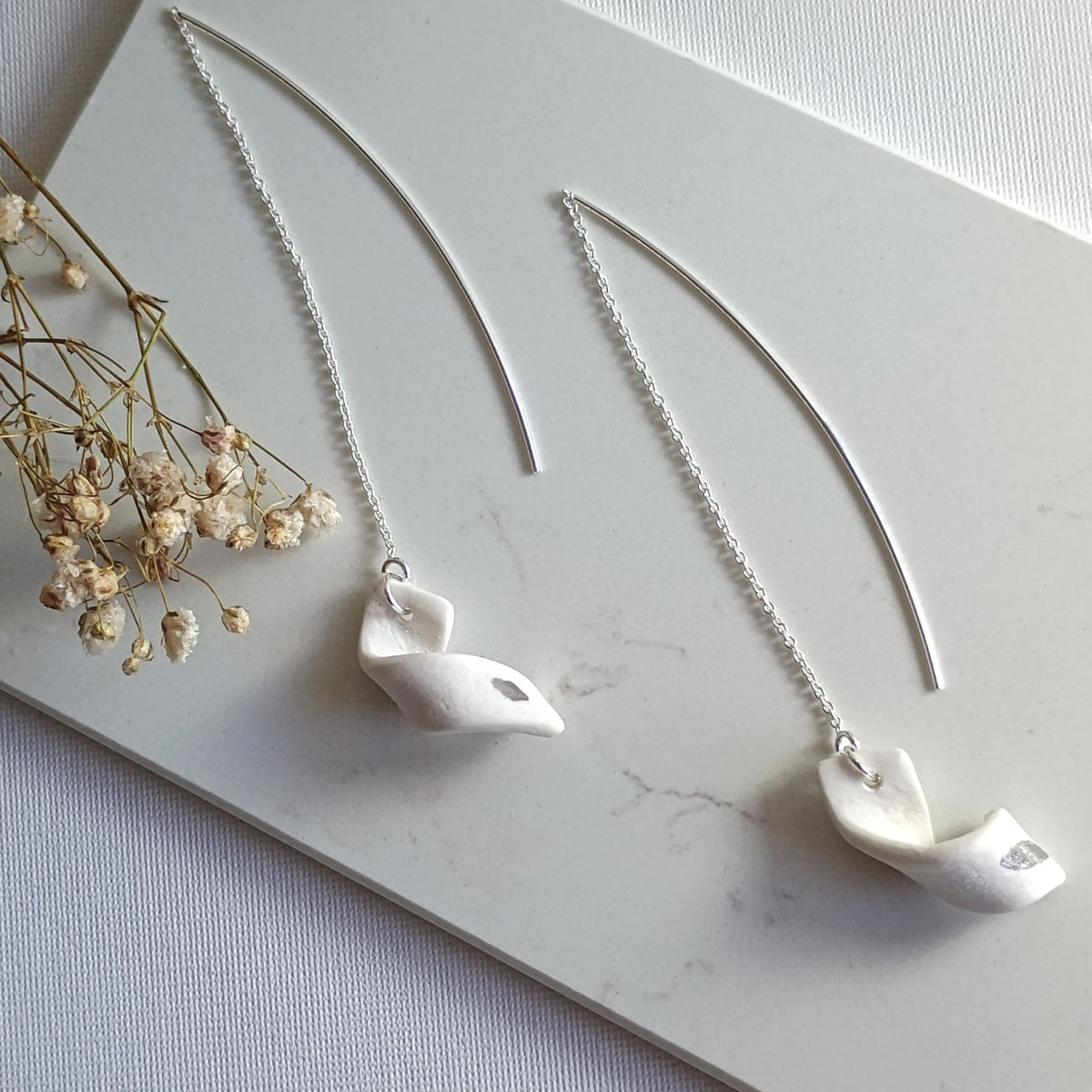 Coppia di orecchini Turbine Maviart in posa dall’alto, porcellana bianca con inclusioni in platino e catenelle in argento, accanto a fiori secchi.