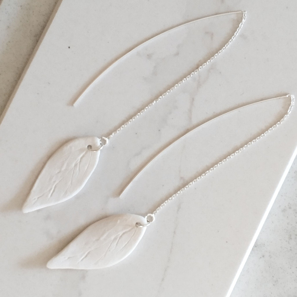 Coppia di orecchini Foglia Maviart in posa dall'alto, foglie in porcellana bianca con venature e catenelle in argento.