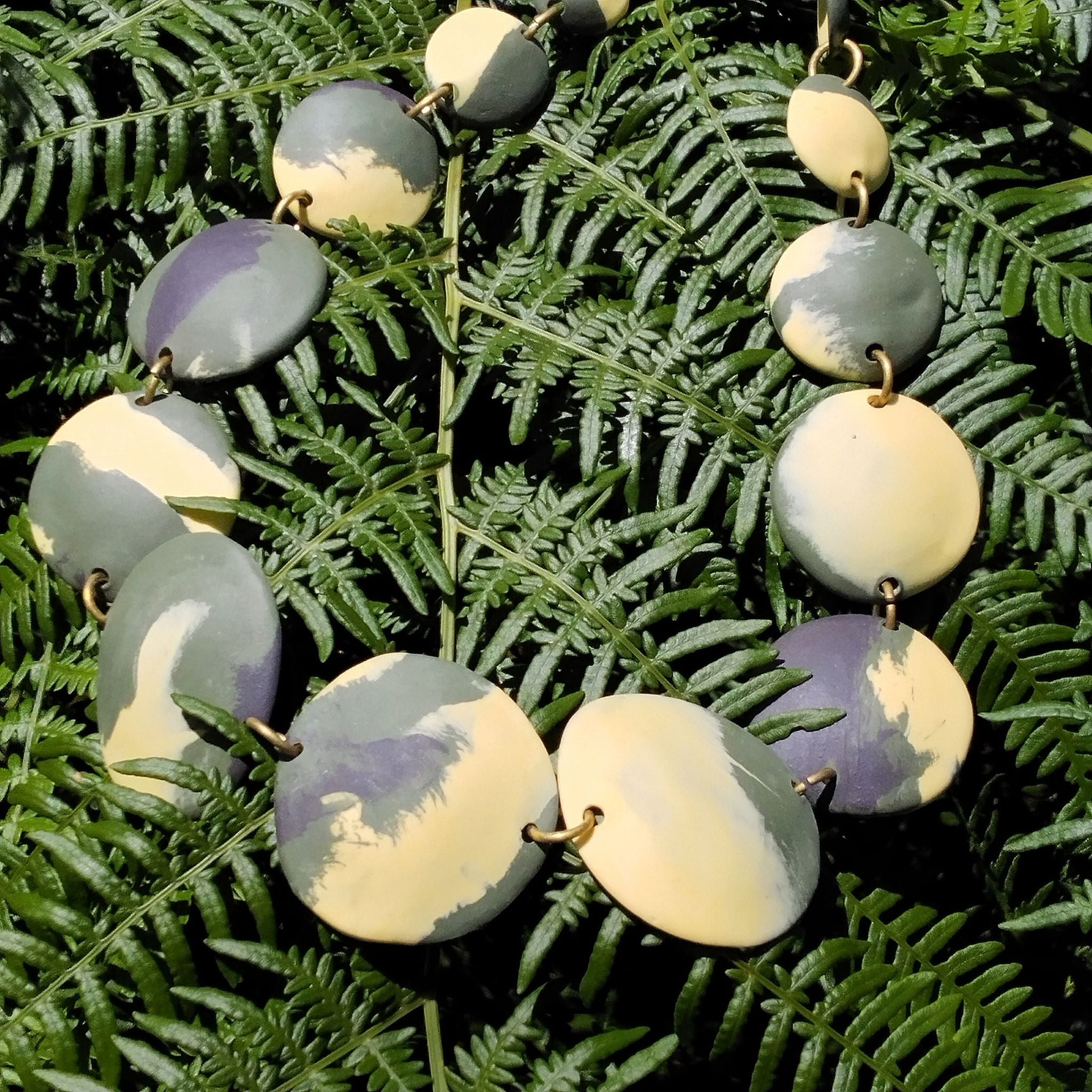 Collana nerikomi porcellana artigianale con elementi convessi grigio-verde e crema, posa dall'alto su felce verde – Maviart